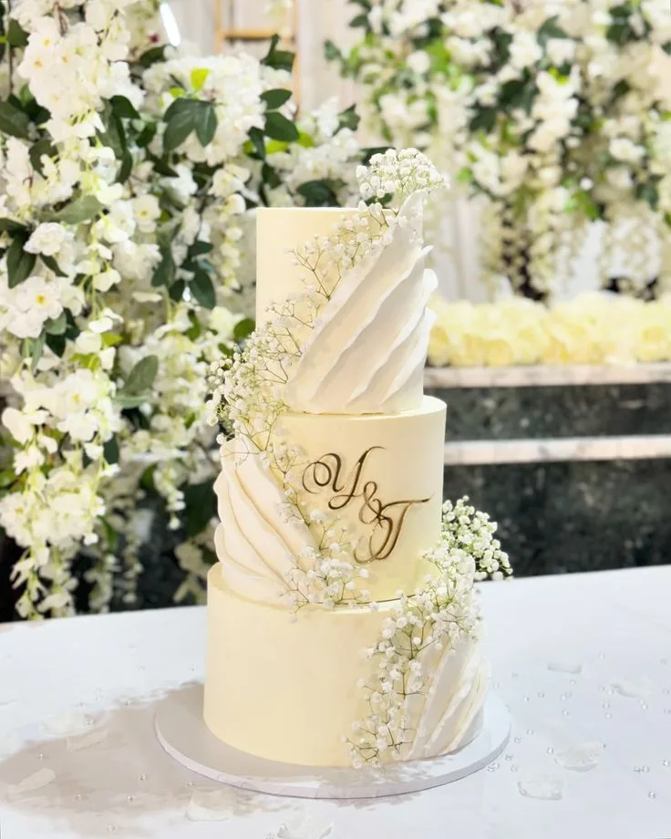 Three-tier ivory wedding cake with fondant drapes, baby's breath and gold monogram by Simply Sweet Udaipur